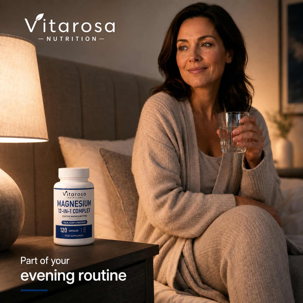 A woman in a cozy bedroom setting holds a glass of water beside a bottle of Vitarosa Nutrition Magnesium 2-in-1 Complex. The scene is warmly lit by a bedside lamp, with the woman wearing a beige cardigan and matching pants.