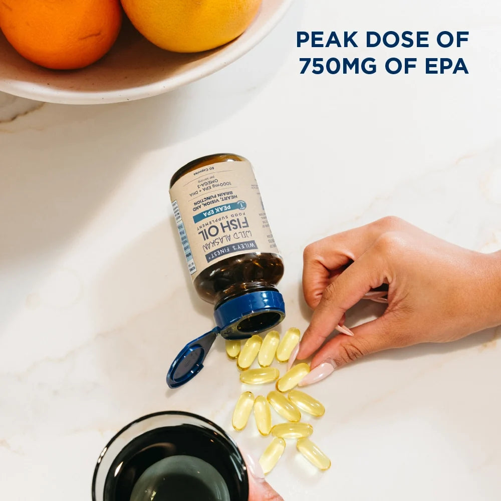 A bottle of fish oil capsules lies on a white marble surface with several yellow softgel capsules spilled out. A hand is reaching toward the capsules, and a bowl of oranges is visible in the background.