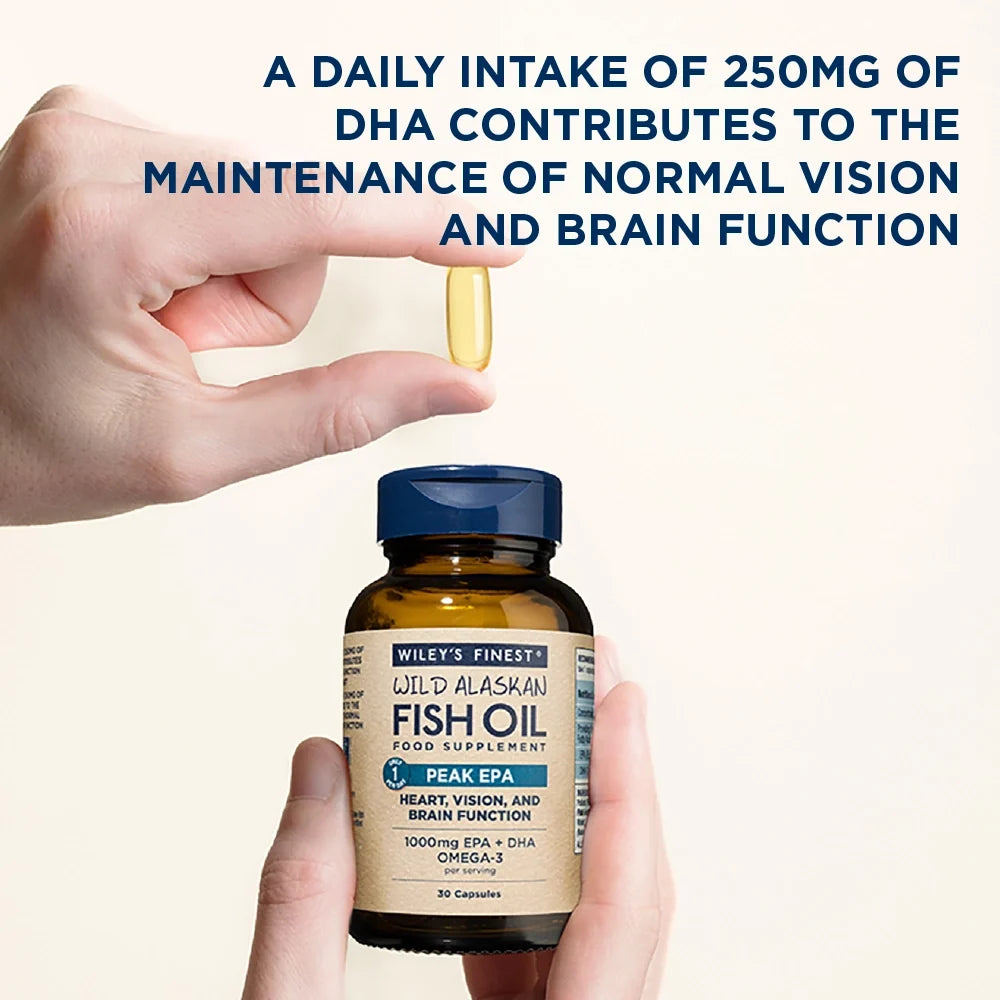 Hand holding a bottle of Wiley's Finest Wild Alaskan Fish Oil with a pill above it, on a beige background.